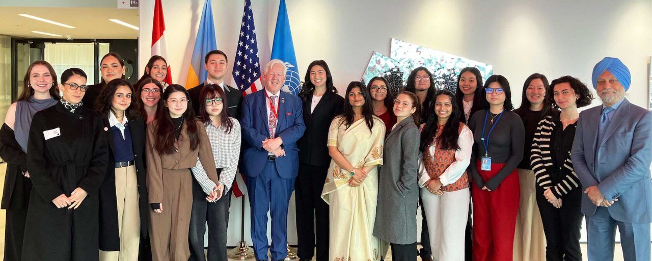 A diverse group of people stands in a brightly lit room, smiling for a photo in front of Canadian, UN, and U.S. flags.