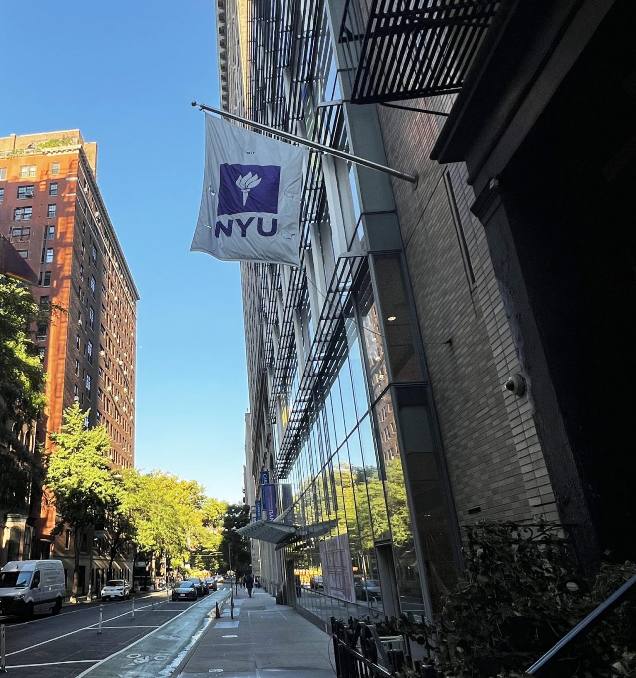 An NYC streetscape taken from the sidewalk, centered on a white and purple NYU flag in front of a building full of gleaming windows.