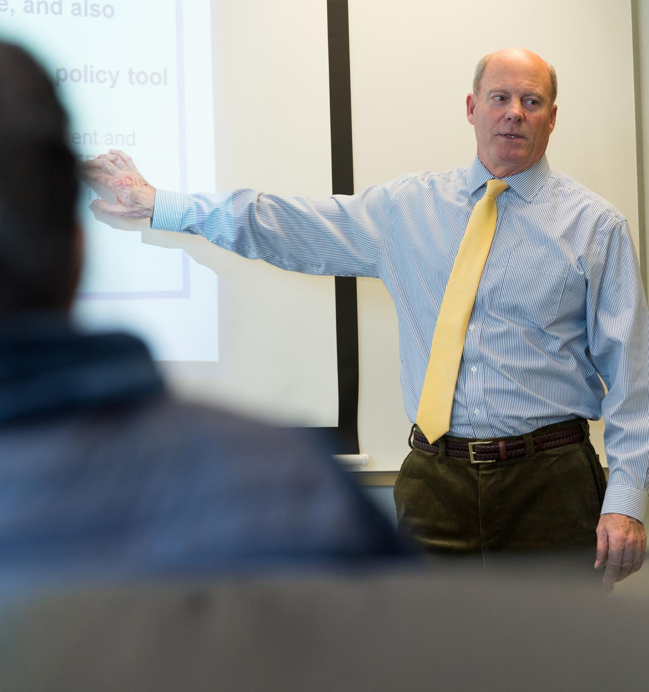 A man wearing a striped shirt and yellow tie gestures towards a projected presentation while speaking to an audience