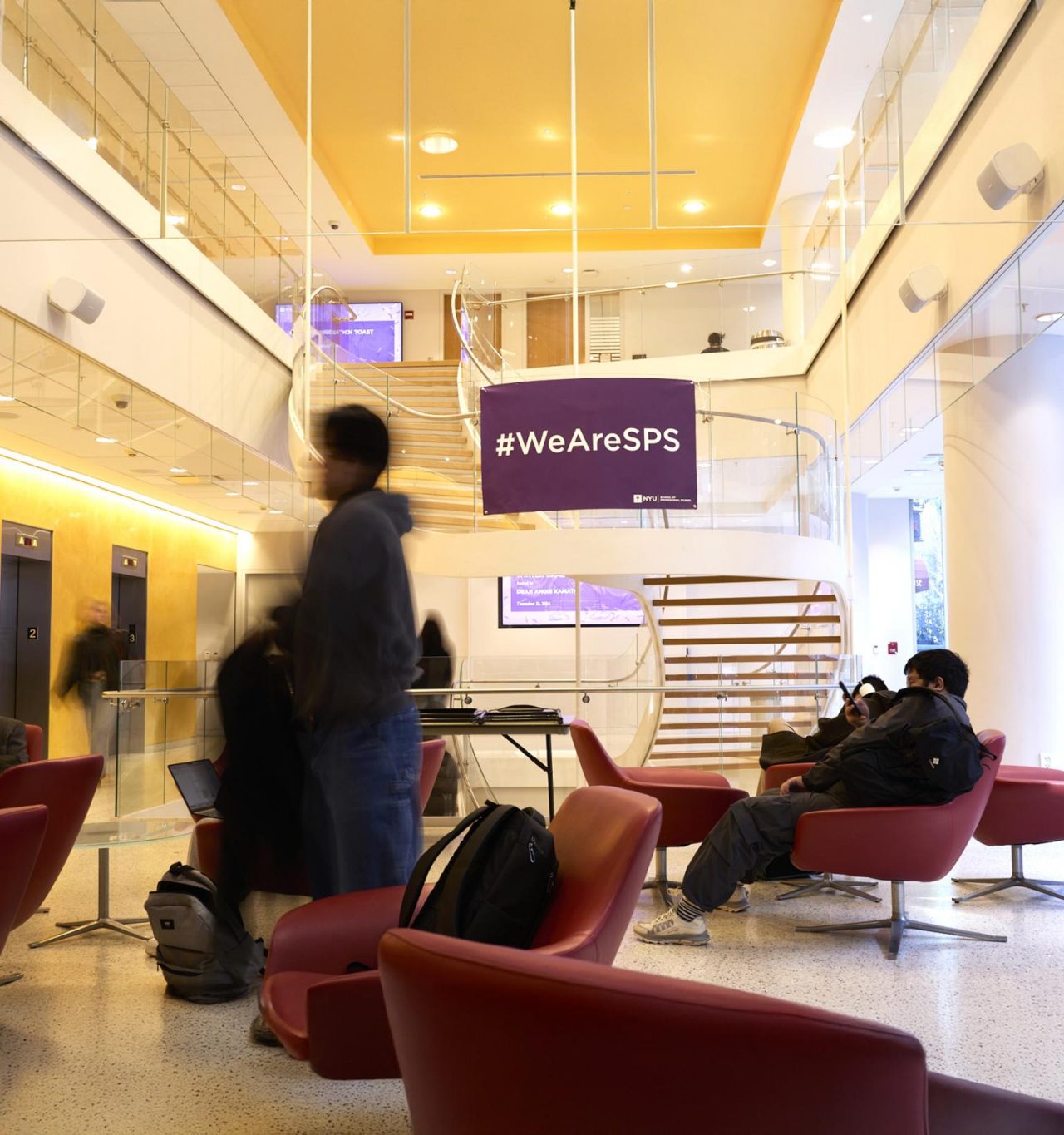 Students mill about or sit in red chairs in an NYU SPS building where a banner reads #WeAreSPS