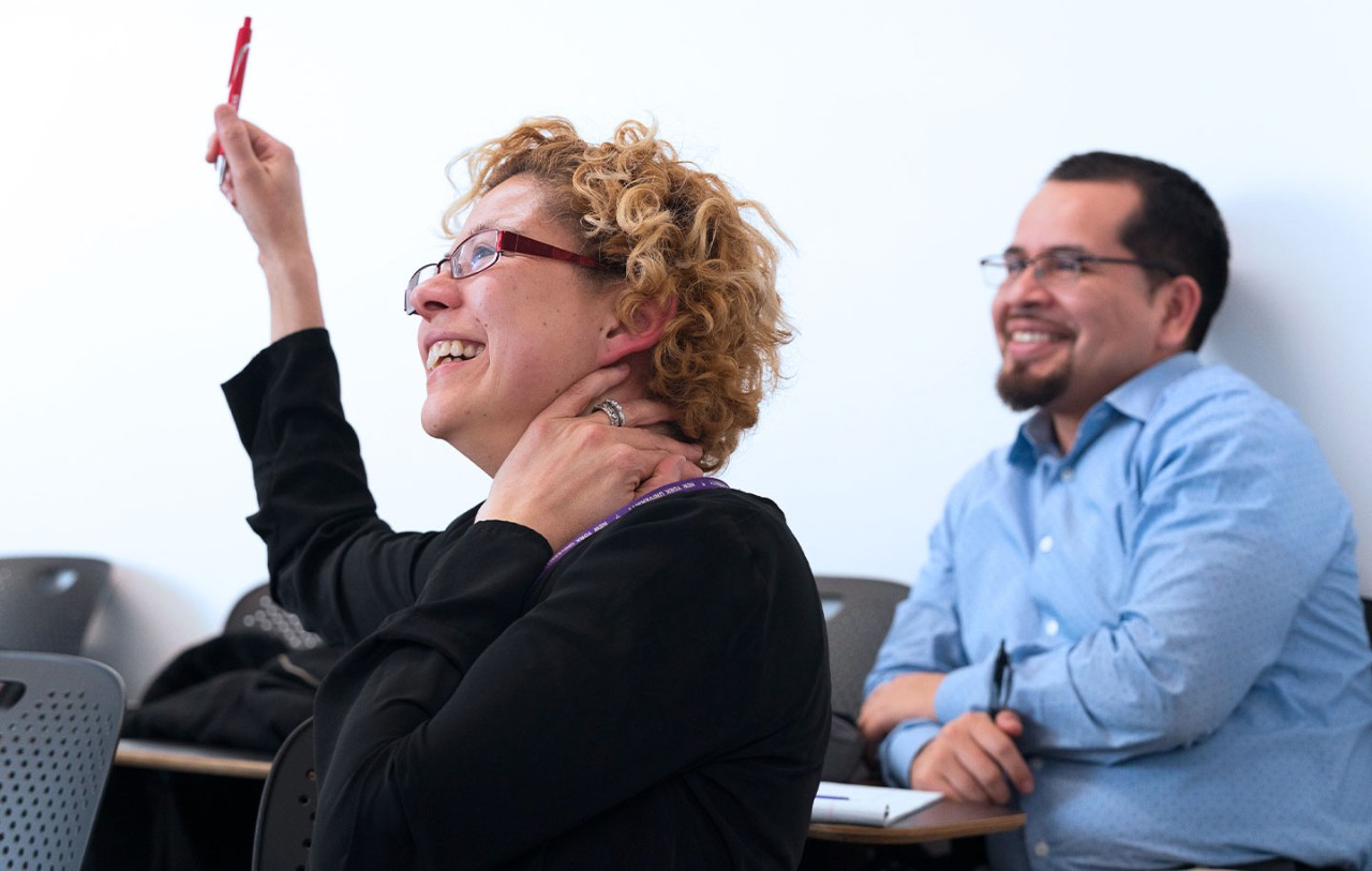 Two continuing education program students at NYU SPS listen to a lecture and one raises her hand