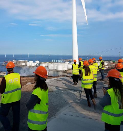 People in safety vests and hard hats near a wind turbine by storage tanks and water.