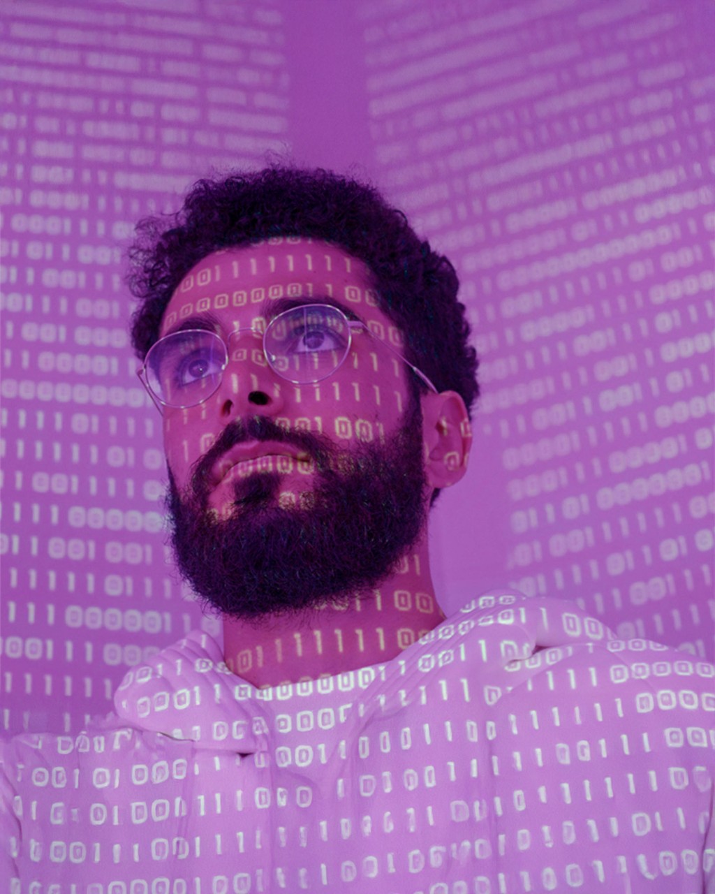 Man with glasses and a beard looks thoughtful as binary code in purple light is projected onto his face, symbolizing technology and cybersecurity
