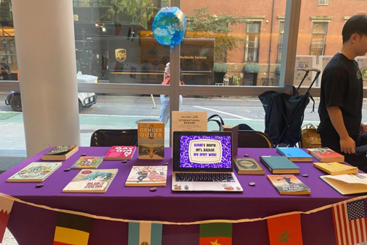 A laptop with an image reading "SCRIBE's Booth Int'l Bazaar SPS Spirit Week" sits on a table with other books in front of a window facing out to a busy NYC street