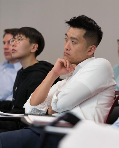 A student in a white shirt sits in a classroom, looking thoughtful, with two others in the background also attentive.