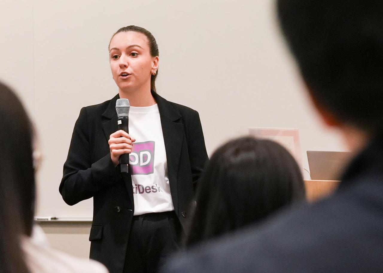 A person speaking in front of a classroom audience with a microphone.