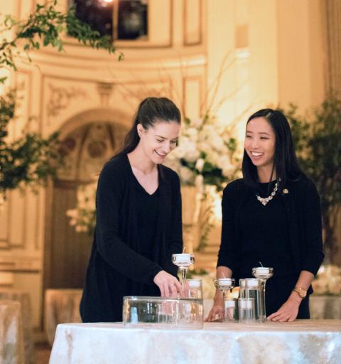 Two women, smiling, stand at a lavishly decorated table in an elegant room with ornate architecture. They're arranging candles.
