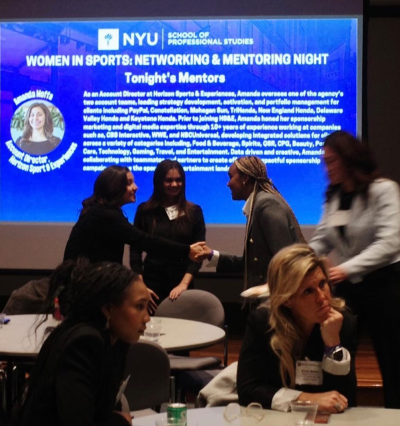 Many women network in a busy ballroom in front of a stage featuring a slide that reads "Women in Sports: Networking and Mentoring Night."