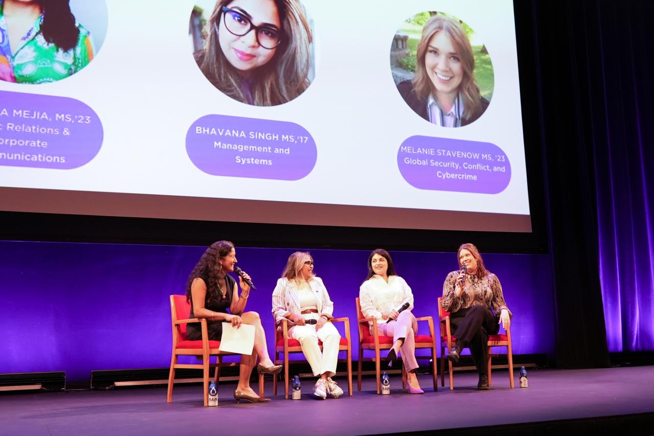 Four women participate in a panel discussion  while seated on-stage in front of a purple backdrop and Powerpoint projection.