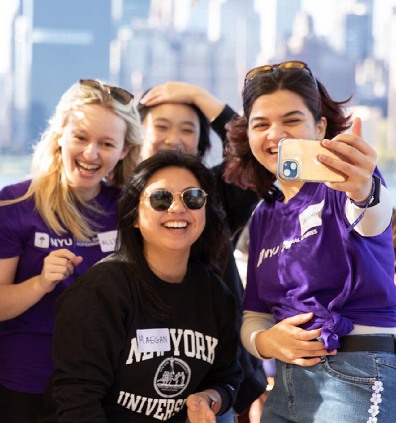 Four NYU SPS students wearing NYU-branded shirts take a selfie while standing in front of a piece of the New York skyline.