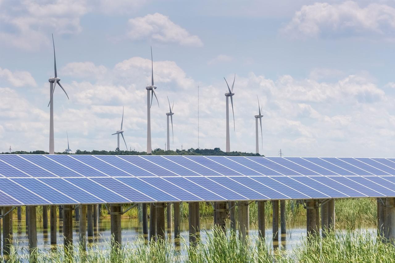 Solar Panels suspended over water in front of a row of wind turbines on a blue, sunny day.