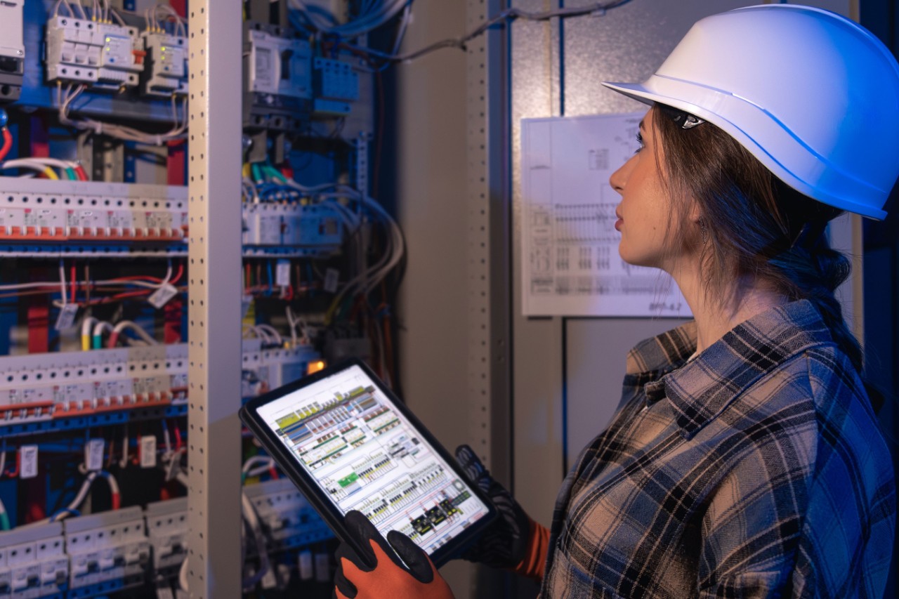 A woman electrician in hardhat inspects switchgear under blue night lighting while reviewing diagrams on a tablet. Visual for smartfactory, automation, iot and safetyfirst in modern industry.