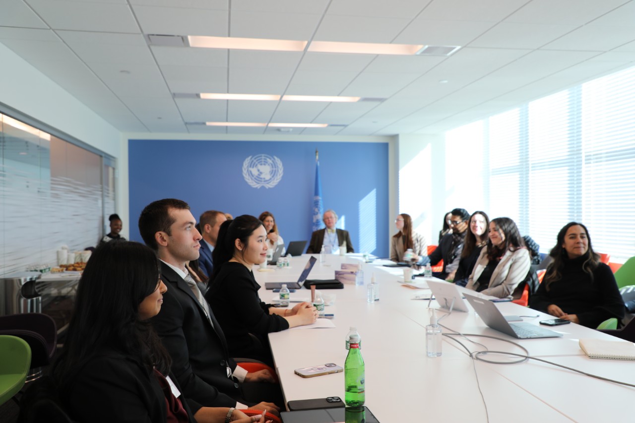 Students and faculty seated around a conference table at the United Nations, listening to a presentation in a bright meeting room with the UN emblem on the wall.