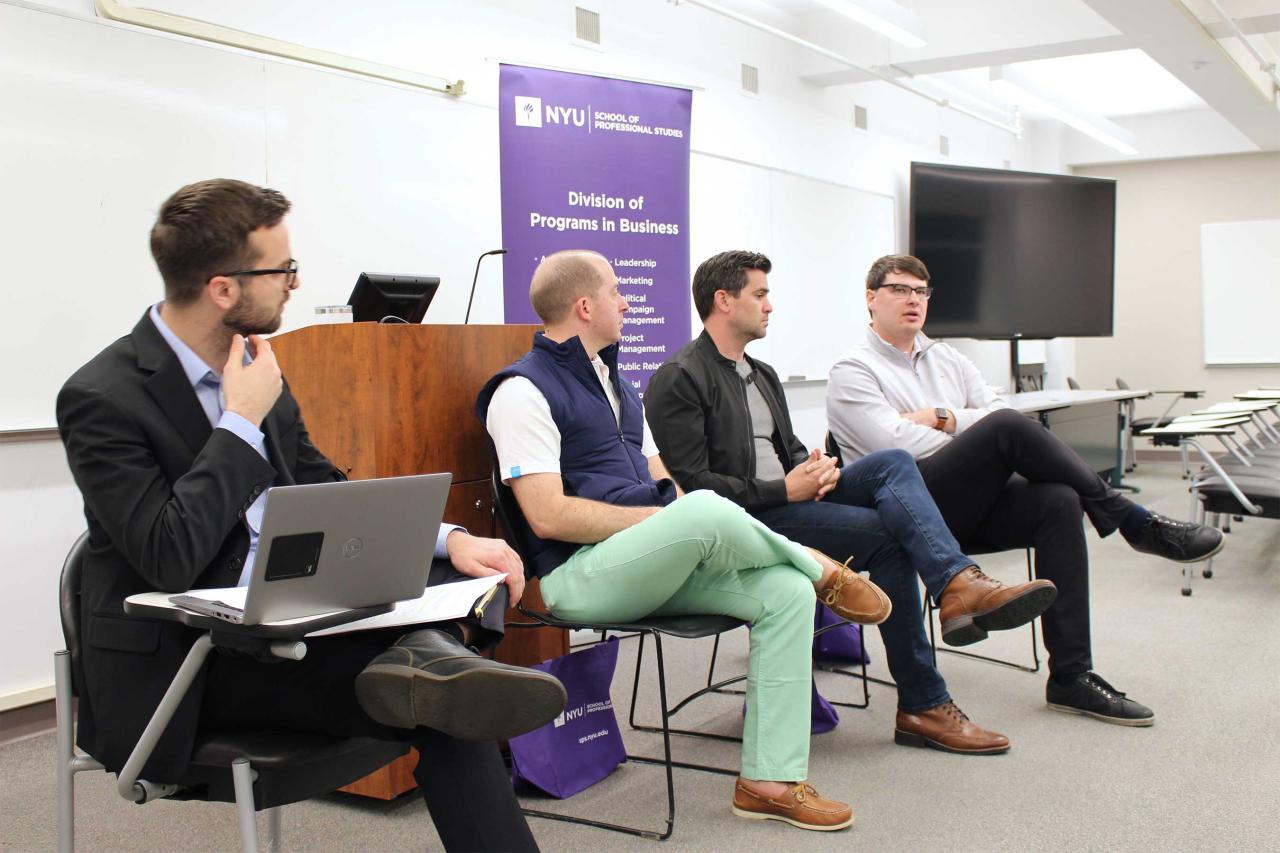 Four men engage in a panel discussion in a classroom. One holds a laptop, others sit casually.