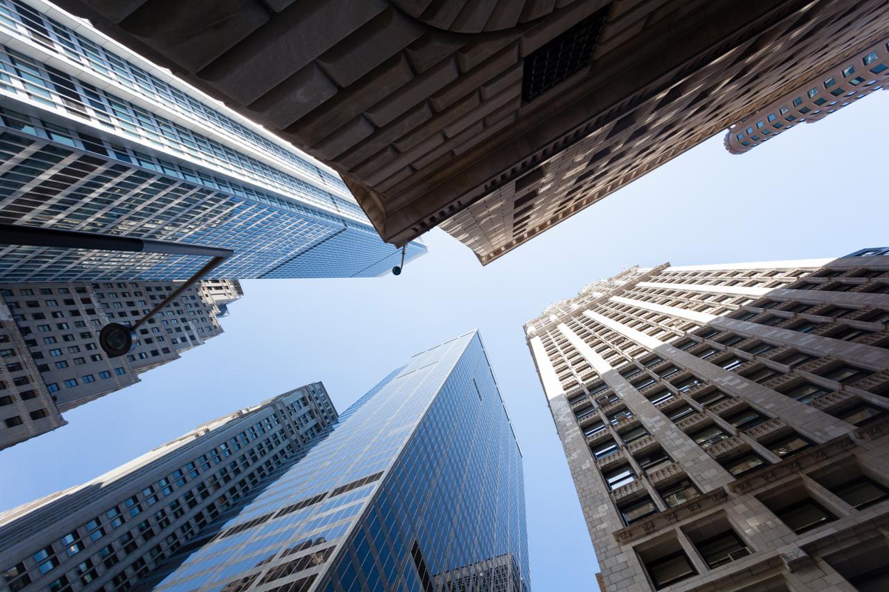 Looking up at skyscrapers from street level, tall buildings with glass and stone facades stretch toward a clear blue sky, conveying urban grandeur.