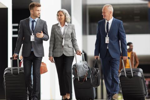 Confident team of forum participants in formalwear carrying wheeled suitcases and crossing airport together of guide