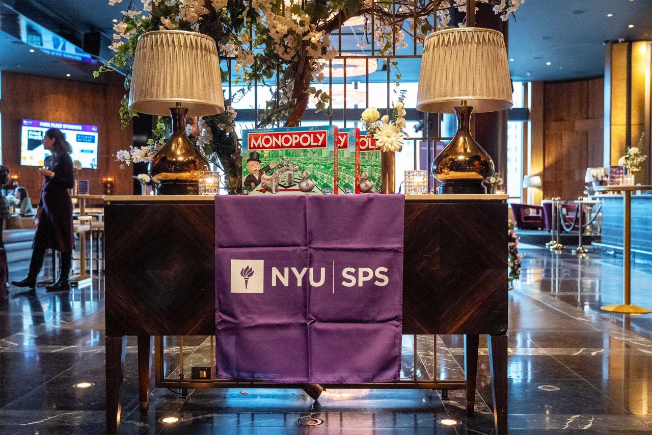 A Monopoly board game displayed on a table with a purple NYU SPS cloth, flanked by two gold table lamps.