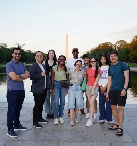 A diverse group of ten people smiles near a reflecting pool with the Washington Monument in the background. The scene is relaxed and cheerful.
