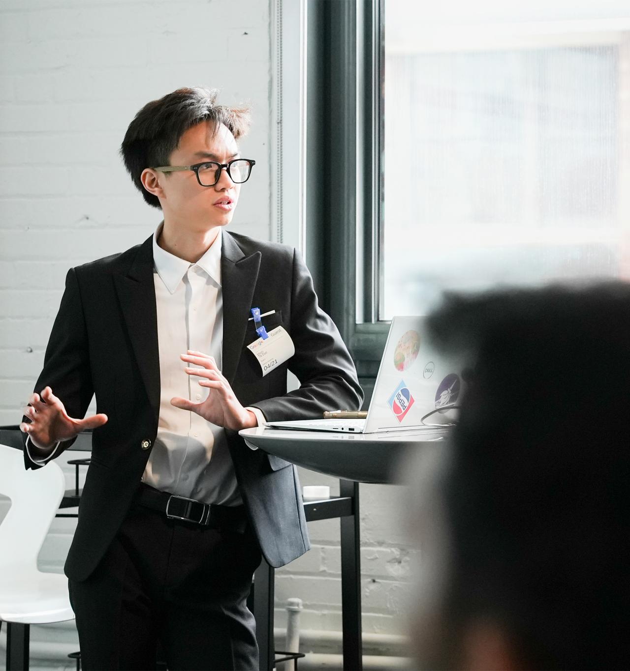 A modern office space with people seated and working. A man A person in a black suit and glasses is speaking passionately in a bright room, standing near a laptop with stickers, conveying a professional setting.    