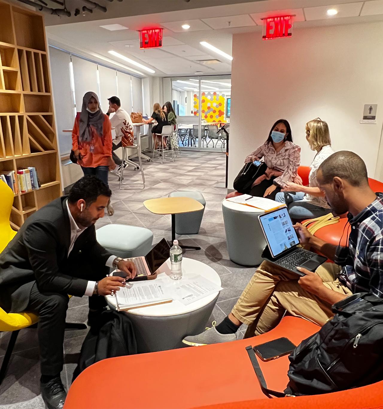 A modern office space with people seated and working. A man in a suit reads papers in a yellow chair, and another man types on a laptop on an orange couch. People converse at tables in the background. The atmosphere is collaborative and focused.