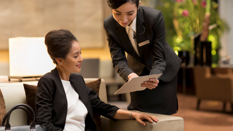 A woman in a black blazer sits on a sofa while a staff member in formal attire offers her a clipboard in a stylish lobby setting.