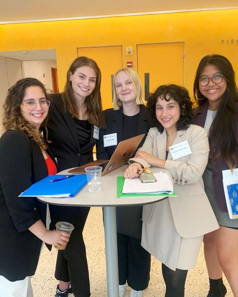 Five women in professional attire smile around a table with a laptop and papers, set against a yellow wall.
