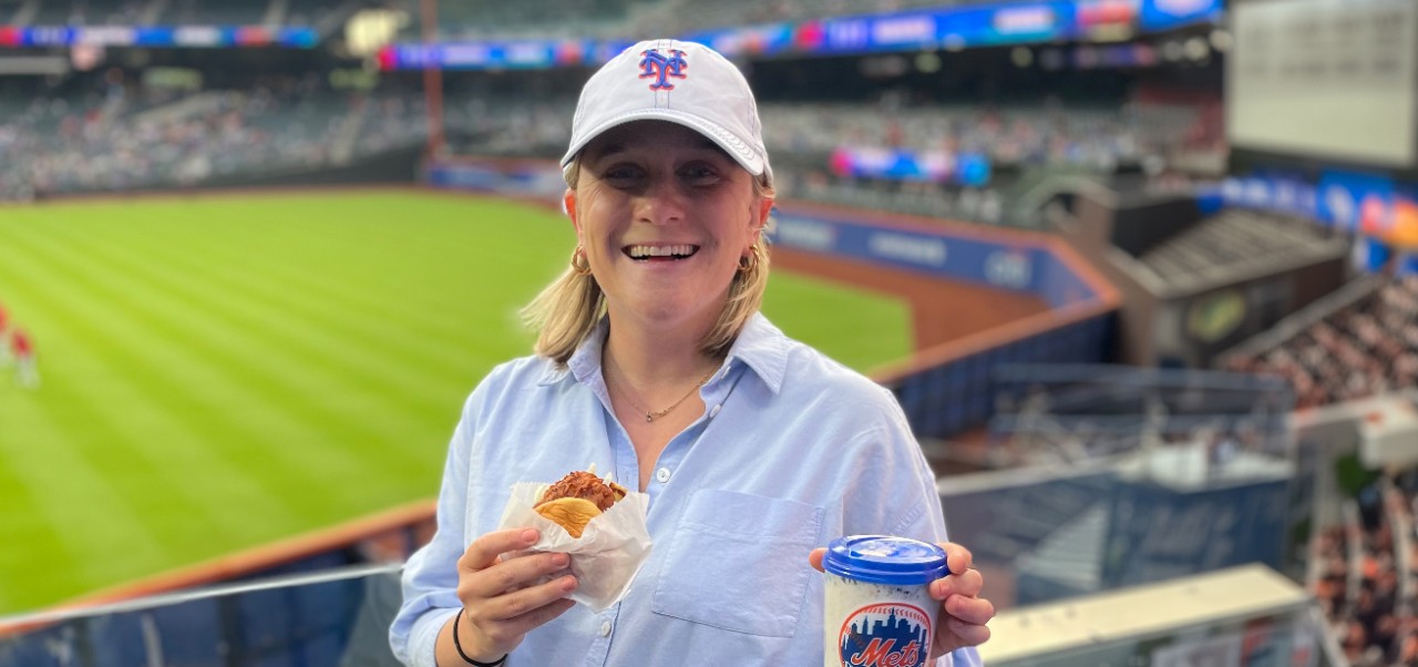 Caroline Kasper, NYU SPS Preston Robert Tisch Institute for Global Sport alumnus, smiles for the camera while holding a sandwich and soft drink at a New York Mets game.