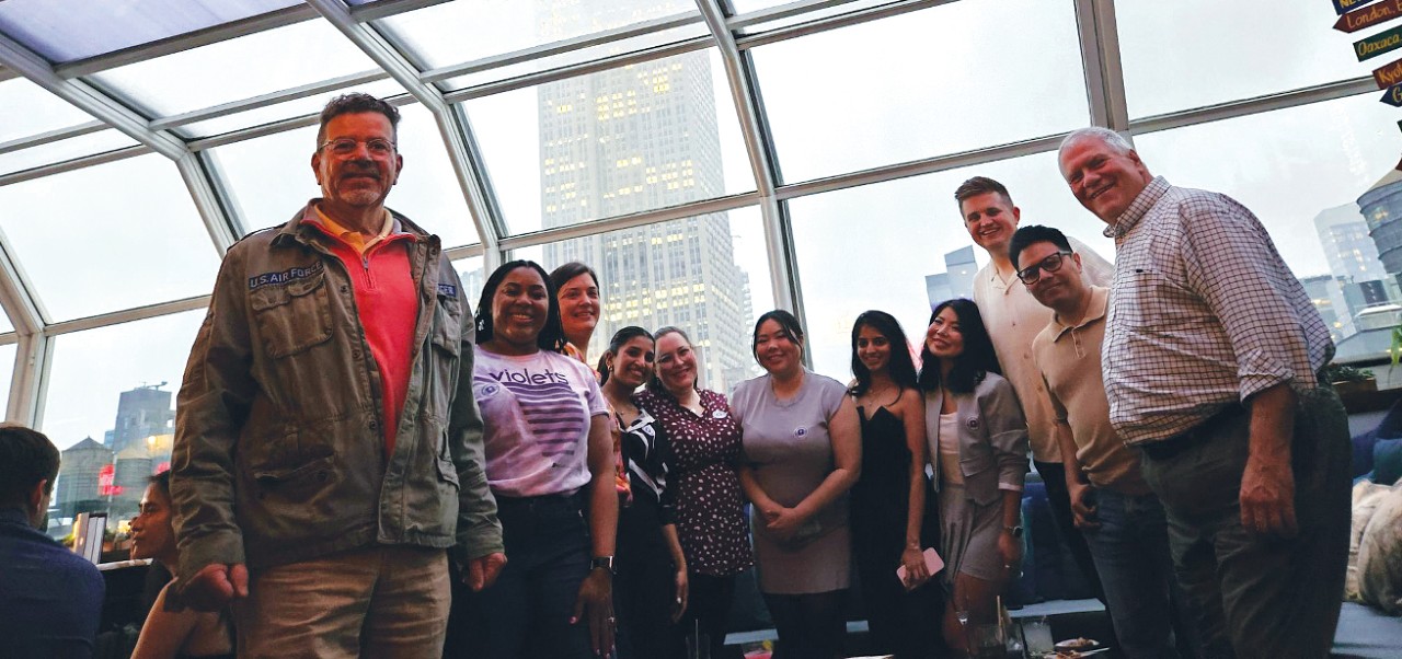 Eva E. K. "EEK" Castleton poses for a photo with members of the NYU SPS community  at a New York restaurant with windowed ceilings.