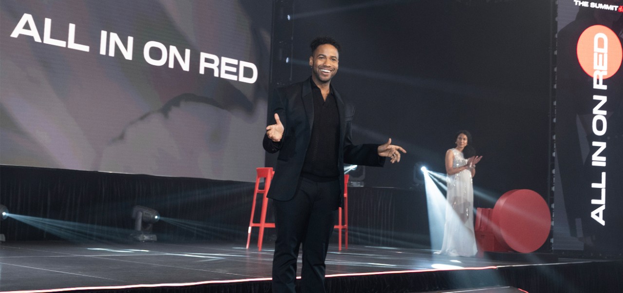 Jeremy Reese, NYU SPS Division of Programs in Business alumnus, clad in a jetblack suit, gives a presentation on a stage in front of a digital banner that reads "All in on Red"