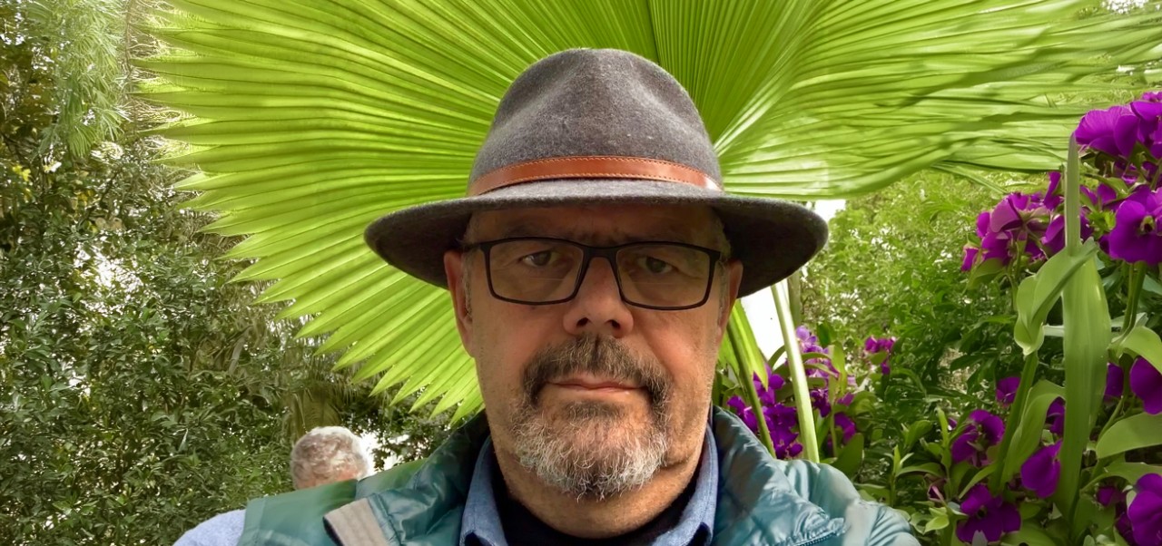 Andres Fortino takes a selfie wearing a grey hat in front of a large palm frond in an outdoor setting with purple flowers.