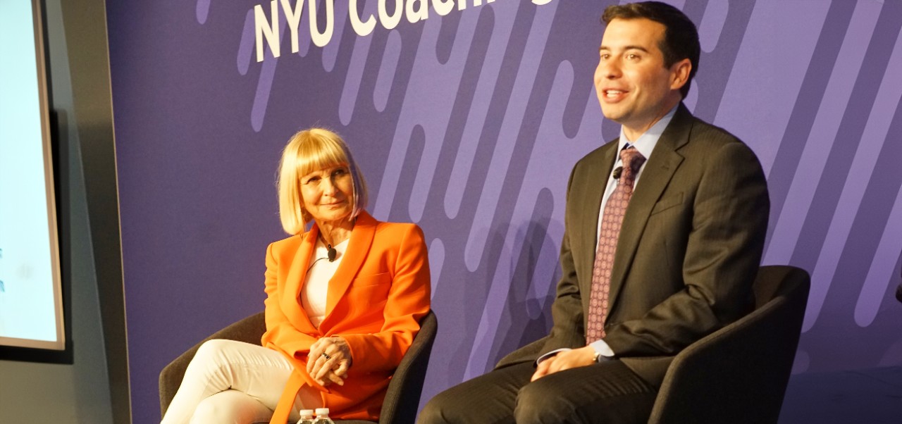 Anna Tavis wears an orange blazer and sits beside Keith Sonderling onstage at NYU SPS's CT Summit with a purple background behind them
