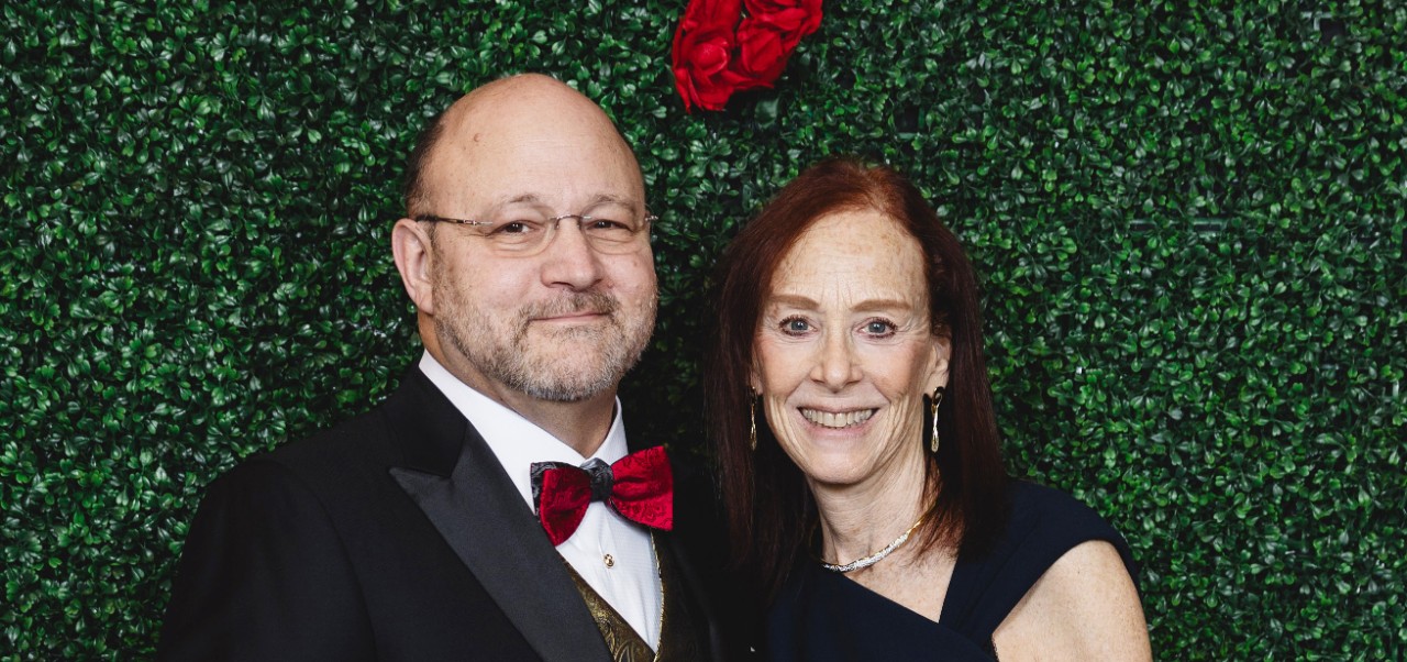 Charles Dorn and his wife pose for a photo at a holiday event at the University of Houston in front of a leafy background