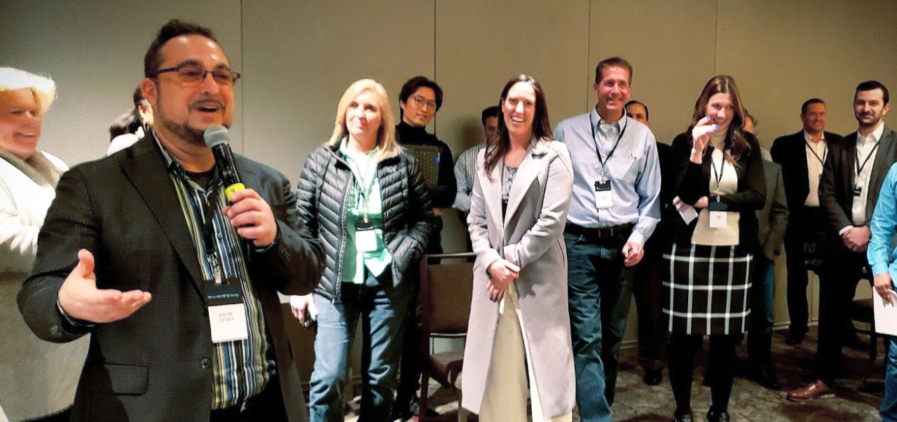 Dave Lipsky speaks into a microphone at an event with about of dozen other professionals listening while standing behind him