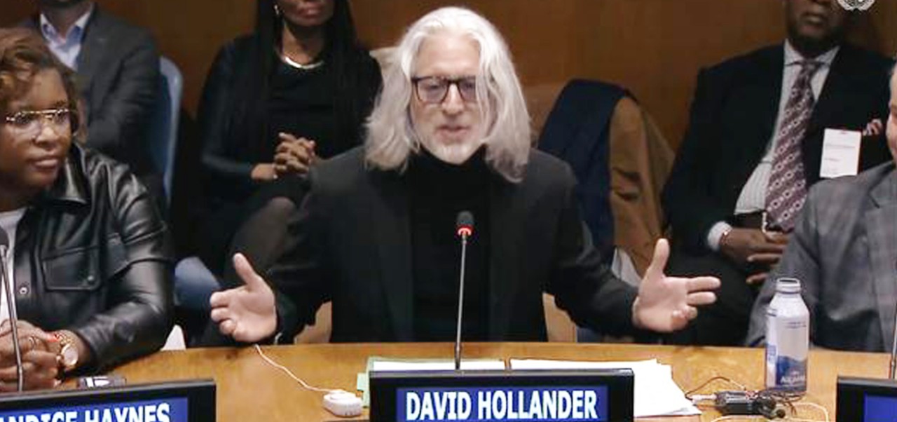 Addressing the U.N. at first every World Basketball Day, David Hollander speaks into a microphone with arms open while sat at a table with his name tag in front of him