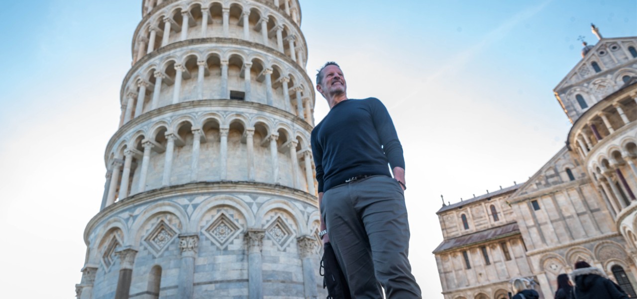 Posing in front of the Tower of Pisa in Italy, Ed Salvato holds a black bag in one hand and has the other hand in his pocket while looking into the distance