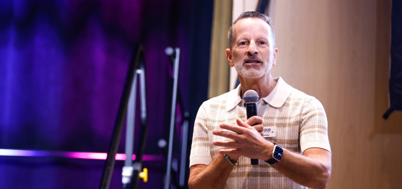 Ed Salvato speaks into a microphone at the Proud Expo 2024 event with a purple and blue-lit stage behind him