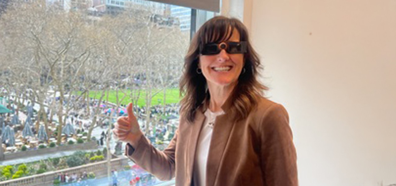 Kathleen Rennie, Clinical Assistant Professor of Marketing and Public Relations, Division of Programs in Business at NYU SPS, gives a "thumbs up" to the camera while standing in front of a window overlooking a park.