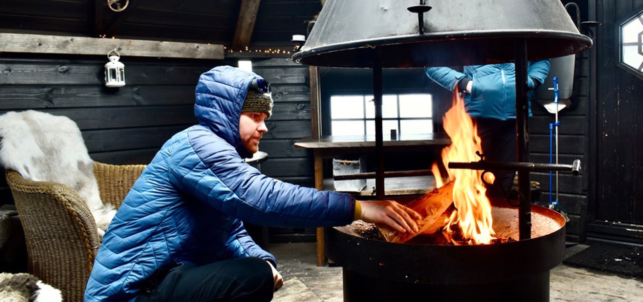 In a cabin in Norway, Nicholas Reese wears a blue jacket while placing wood in a fire pit in the middle of the room