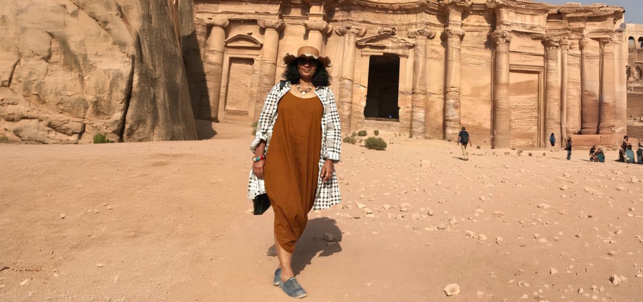 Rajamma Krishnamurthy wears a hat and sunglasses while walking in front of a red stone building in Petra, Jordan