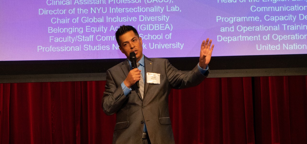 Raul Sanchez stands on a stage with a red curtain and a screen behind him at the NYU SPS Innovation Day event