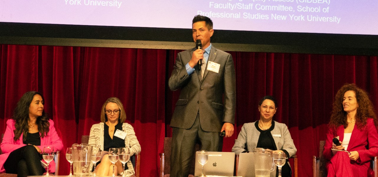 Raul Sanchez stands on a stage with a red curtain  and four women sitting in chairs behind him at the NYU SPS Innovation Day event