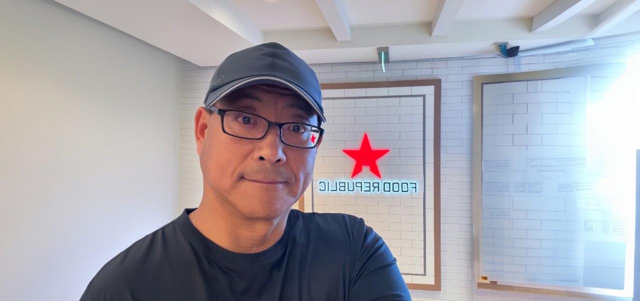 Steven Pae poses for a selfie wearing a baseball cap and a black shirt in front of a Food Republic sign on a white wall
