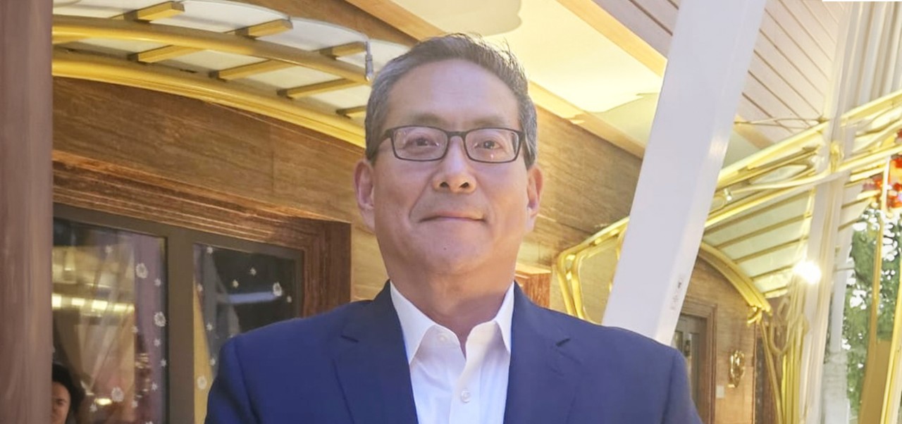 Steven Pae wears a navy suit jacket and a white shirt and smiles in front of a gold and brown building