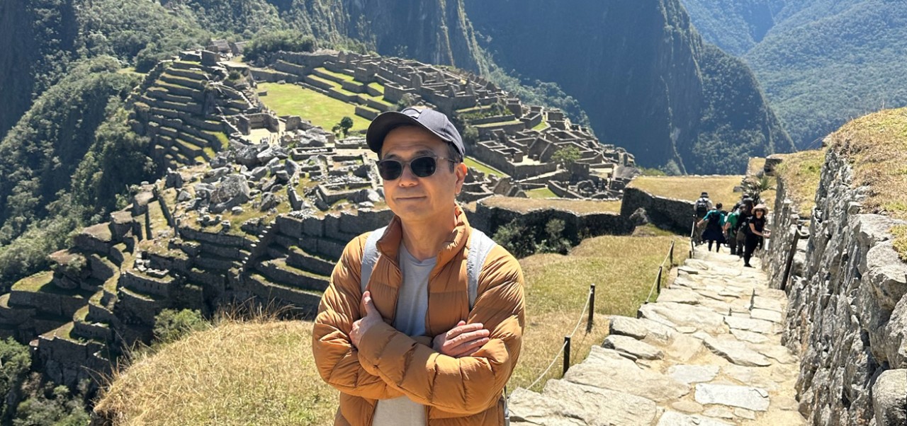 With the ruins of Machu Picchu behind him, Toshi Mitsudomee poses for a picture with his arms crossed and wearing a hat and sunglasses