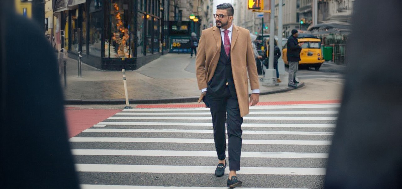 Zair Cheema looks to his right while walking across a crosswalk in London while wearing a tan jacket over a black suiit.