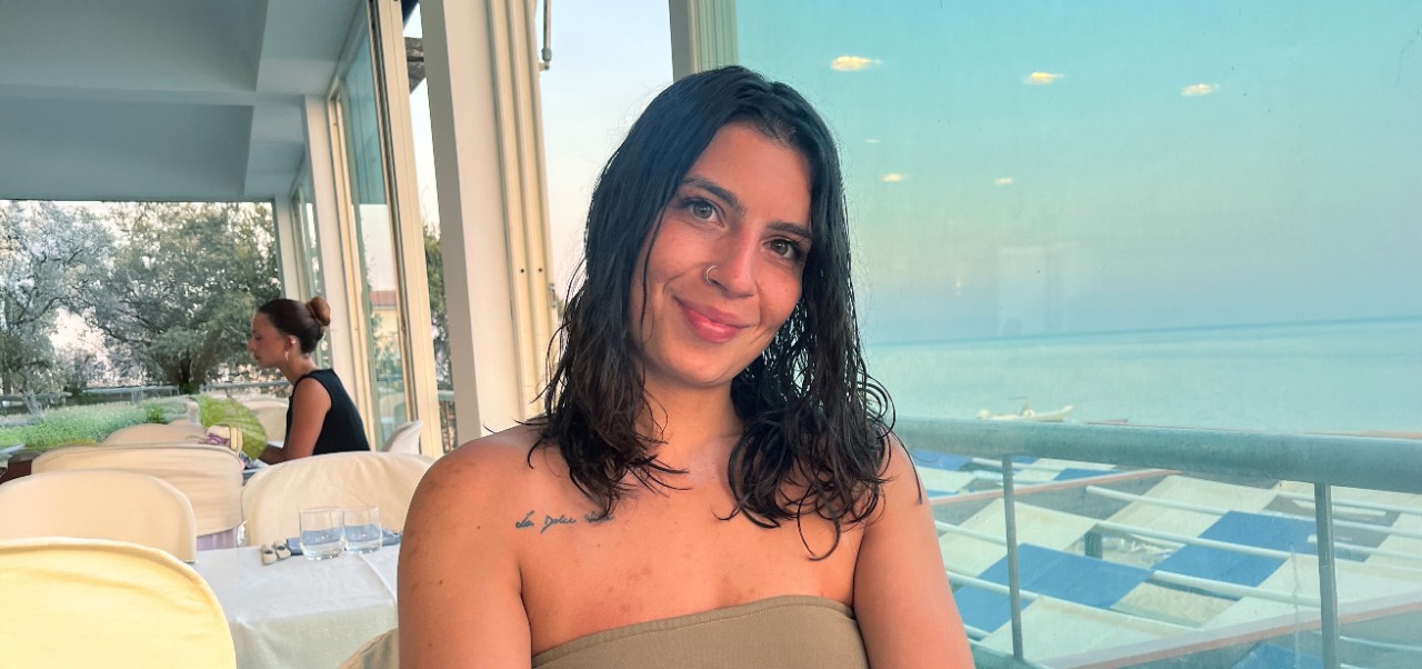 Francesca Dell'Acaqua, student at the Center for Publishing and Liberal Arts, NYU SPS smiles for the camera at a cafe overlooking a body of water.