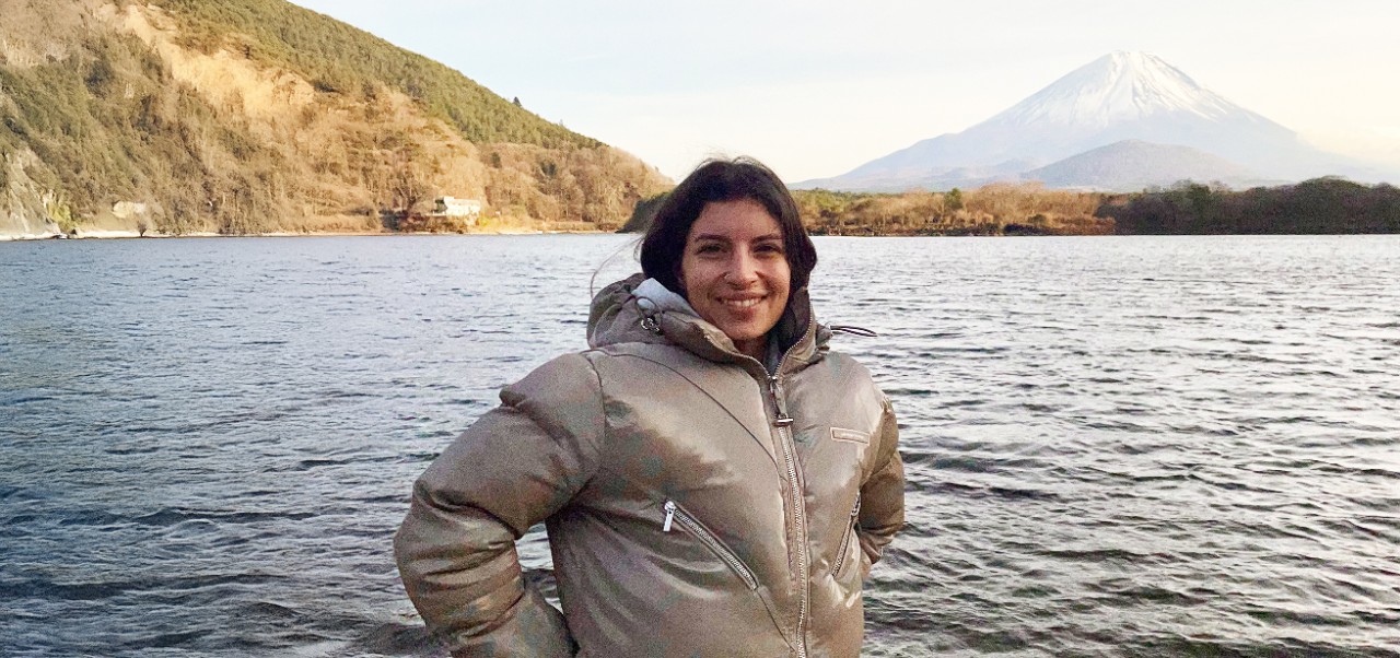 Francesca Dell'Acaqua, student at the Center for Publishing and Liberal Arts, NYU SPS smiles for the camera standing in front of a lake with an imposing mountain in the background.