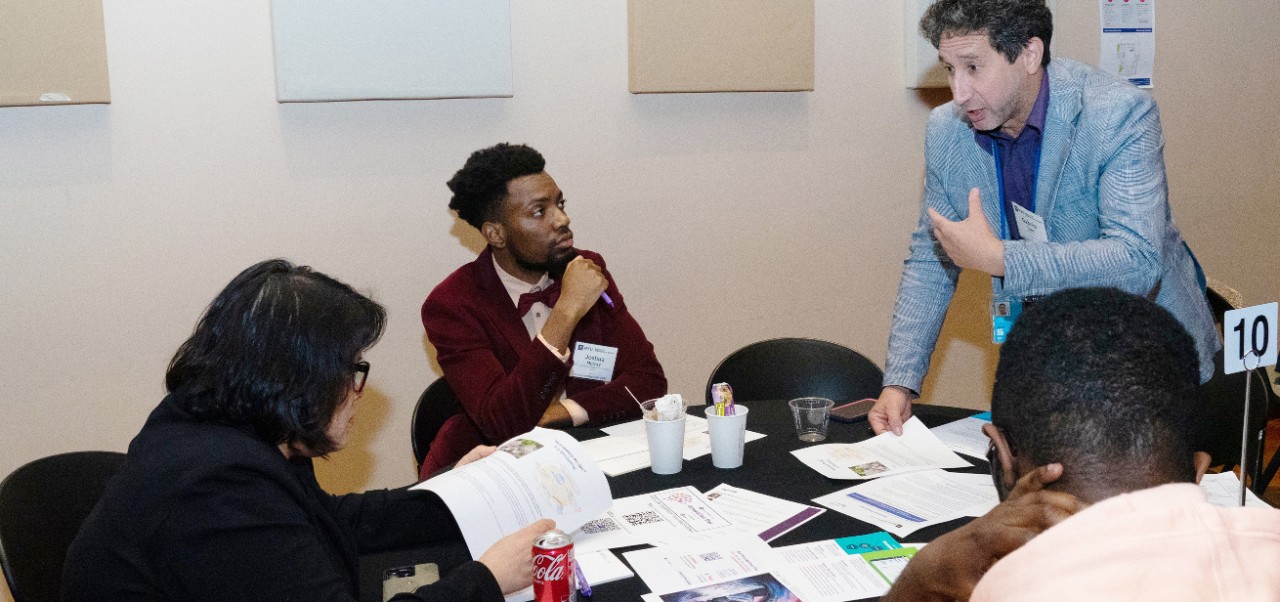  Joshua Murray, Division of Programs in Business student at NYU SPS, works at a cluttered table surrounded by colleagues in a conference room.