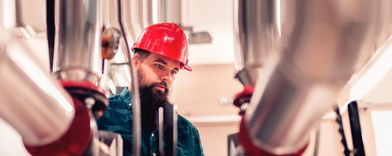 A plumber wearing a red hard hat examines steel pipes.