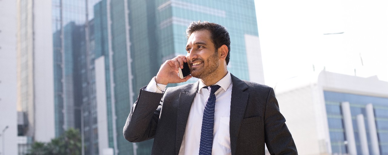 A businessman talks on an iPhone in the middle of an urban streetscape on a sunny day.